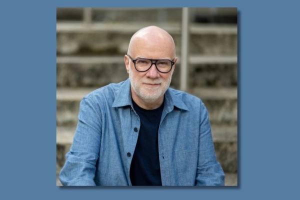 A colour portrait of John Spencer Williams, a white man with grey beard and glasses, who is wearing a blue shirt.